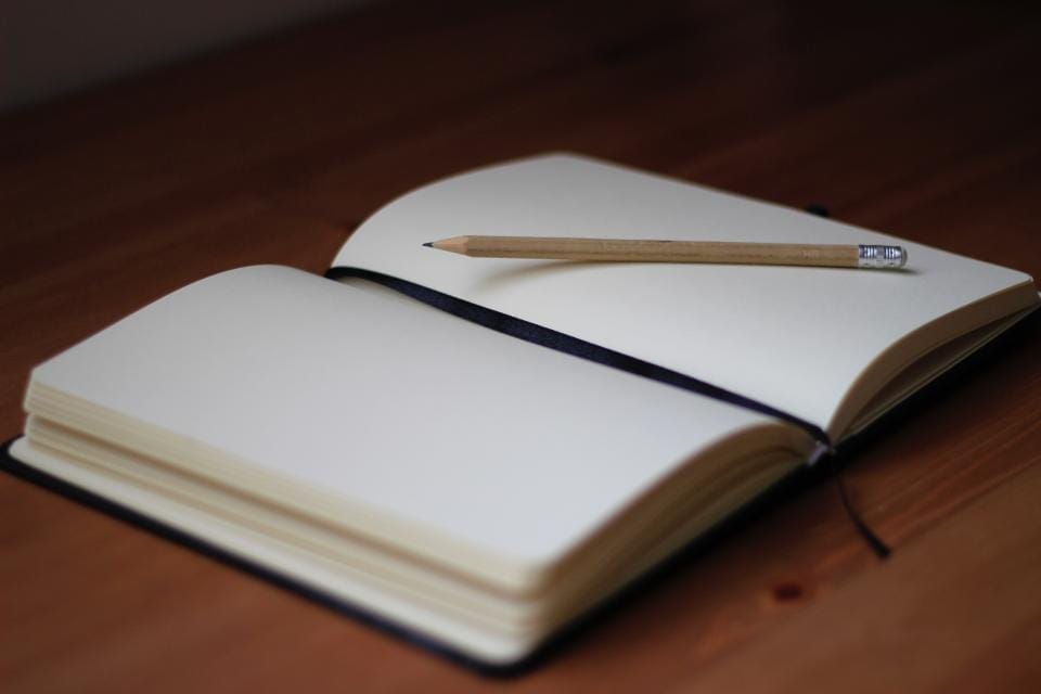 Ahşap bir masanın üzerinde açık duran boş bir defter, sayfanın üstünde bir kurşun kalem